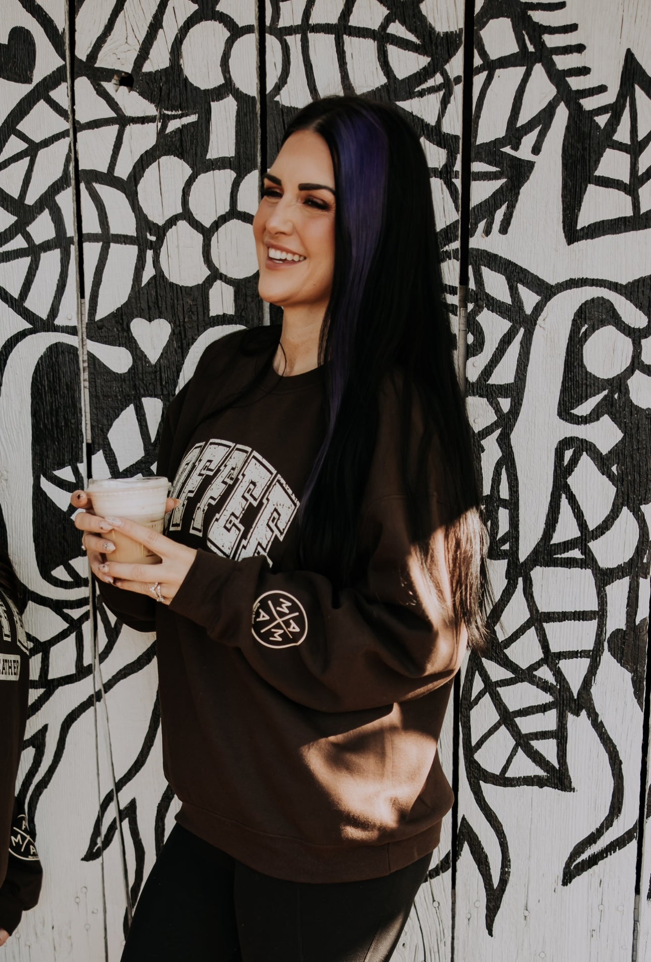 A woman with long dark hair and a purple streak wears the cozy Coffee Weather Crewneck by Mama X™ Brand, smiling with a drink in hand as she stands before an abstract black and white mural.