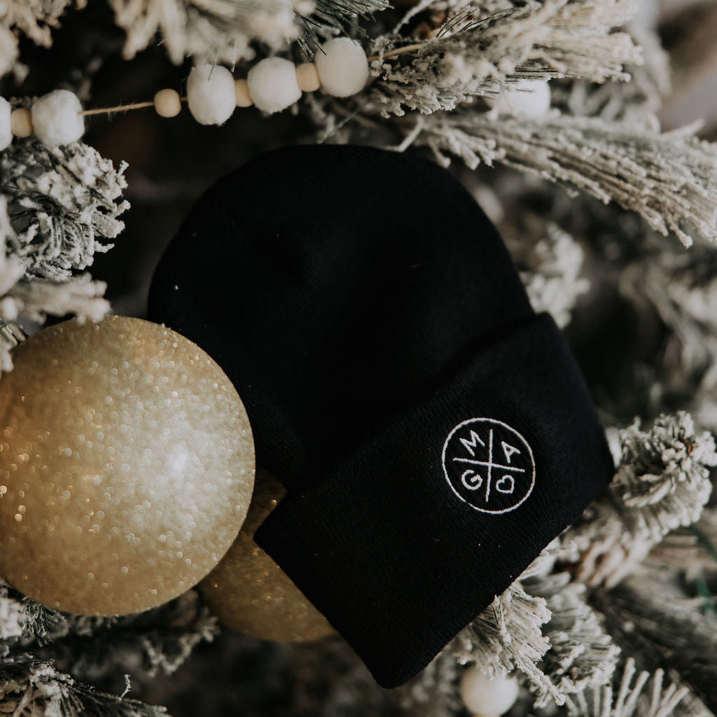 A cozy GMA Beanie by Mama X™ Brand, featuring a white circular logo, hangs on a snow-dusted Christmas tree decorated with gold ornaments and white bead garlands.
