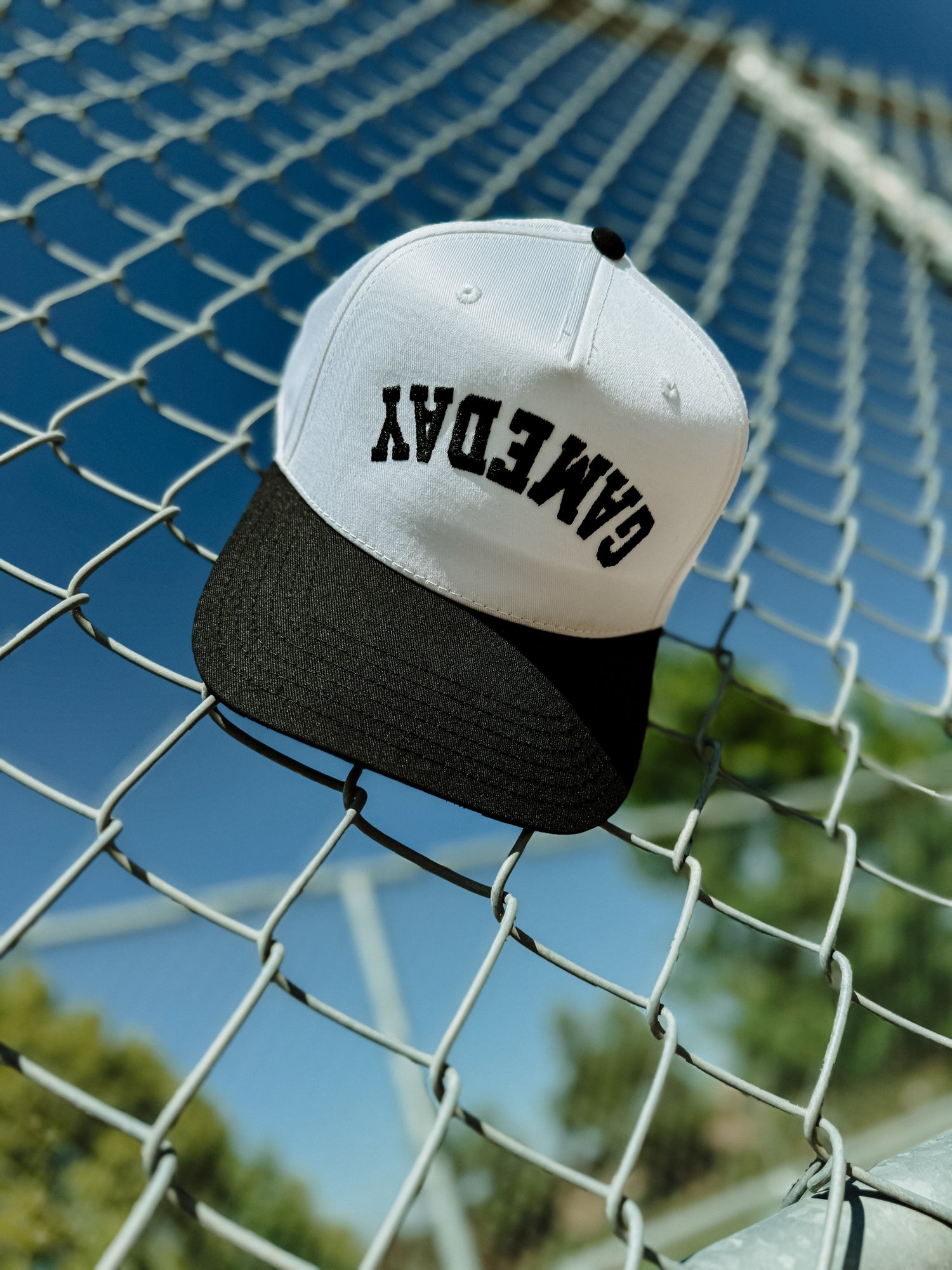 The Mama X™ Brand Game Day Trucker—black and white with “Game Day” upside down on the front—rests on a chain-link fence beneath a sunny sky, with blurred trees and a sports field in the background.