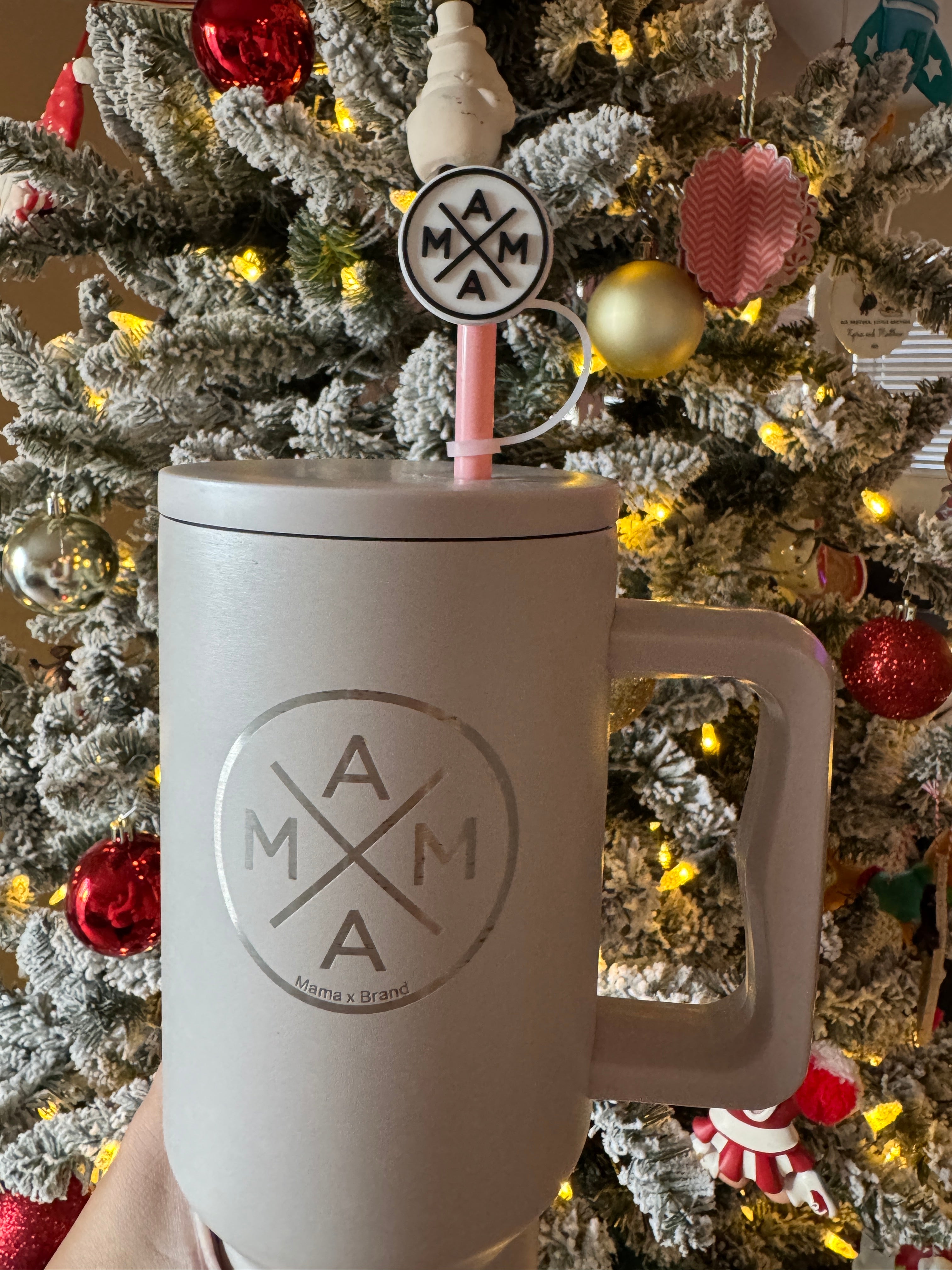 A hand holds a large, two-toned Mama X™ Brand mug featuring the "MAMA" logo. Inside, a Mama X™ Straw Topper with the same logo complements the look. In the background, a decorated Christmas tree adorned with twinkling ornaments and lights adds to the festive scene.