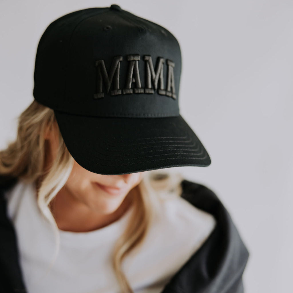 A person with long blonde hair wears the Mama X™ Brand Mama black on black Trucker Hat with 3D embroidery. Their face is partly hidden by the cap, paired with a white shirt and black jacket over their shoulders against a plain background.