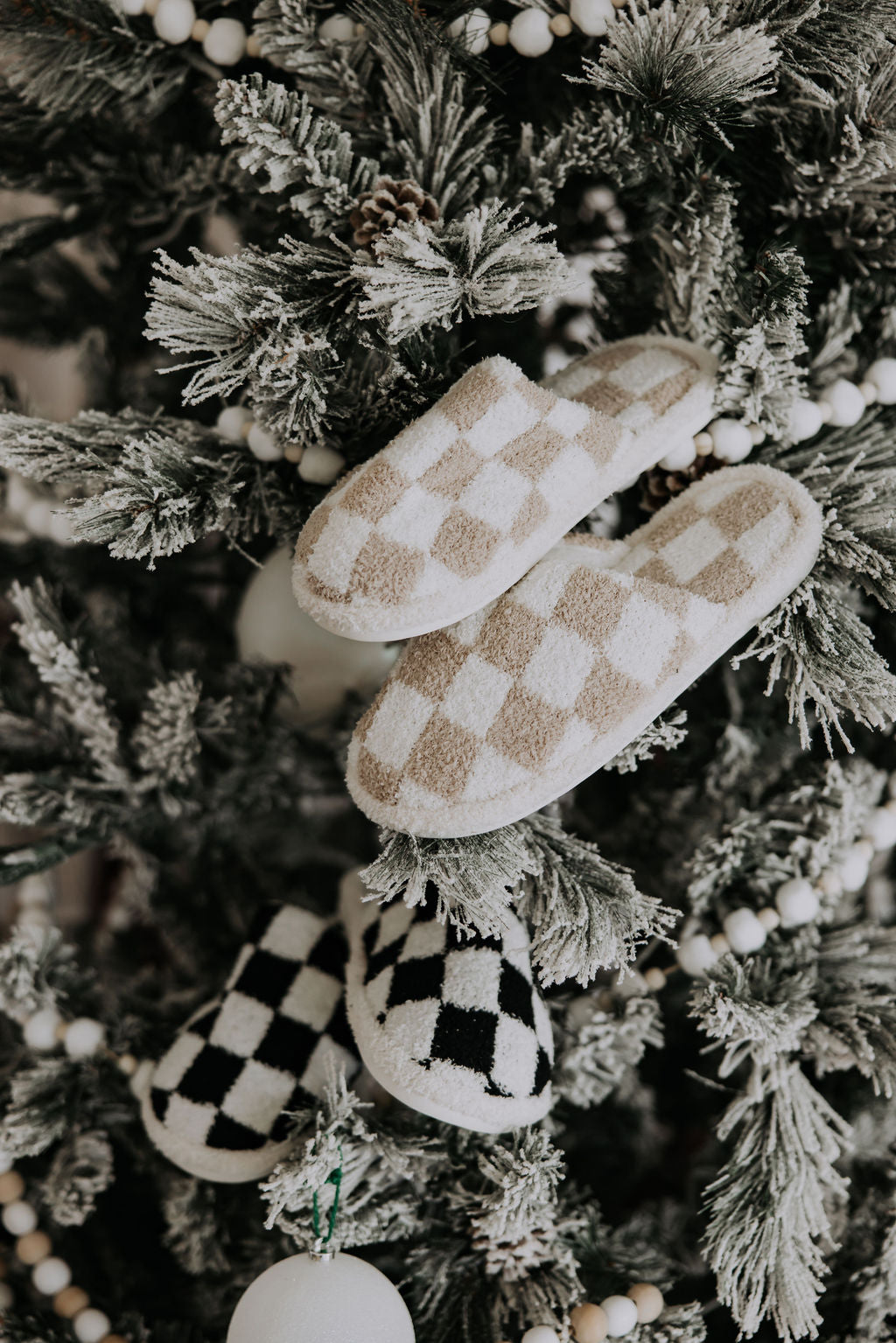 A frosted Christmas tree decorated with Mama X™ Brand's cozy beige and black checkered slippers, featuring the Mama X logo, alongside round white ornaments, creates a warm and festive ambiance.