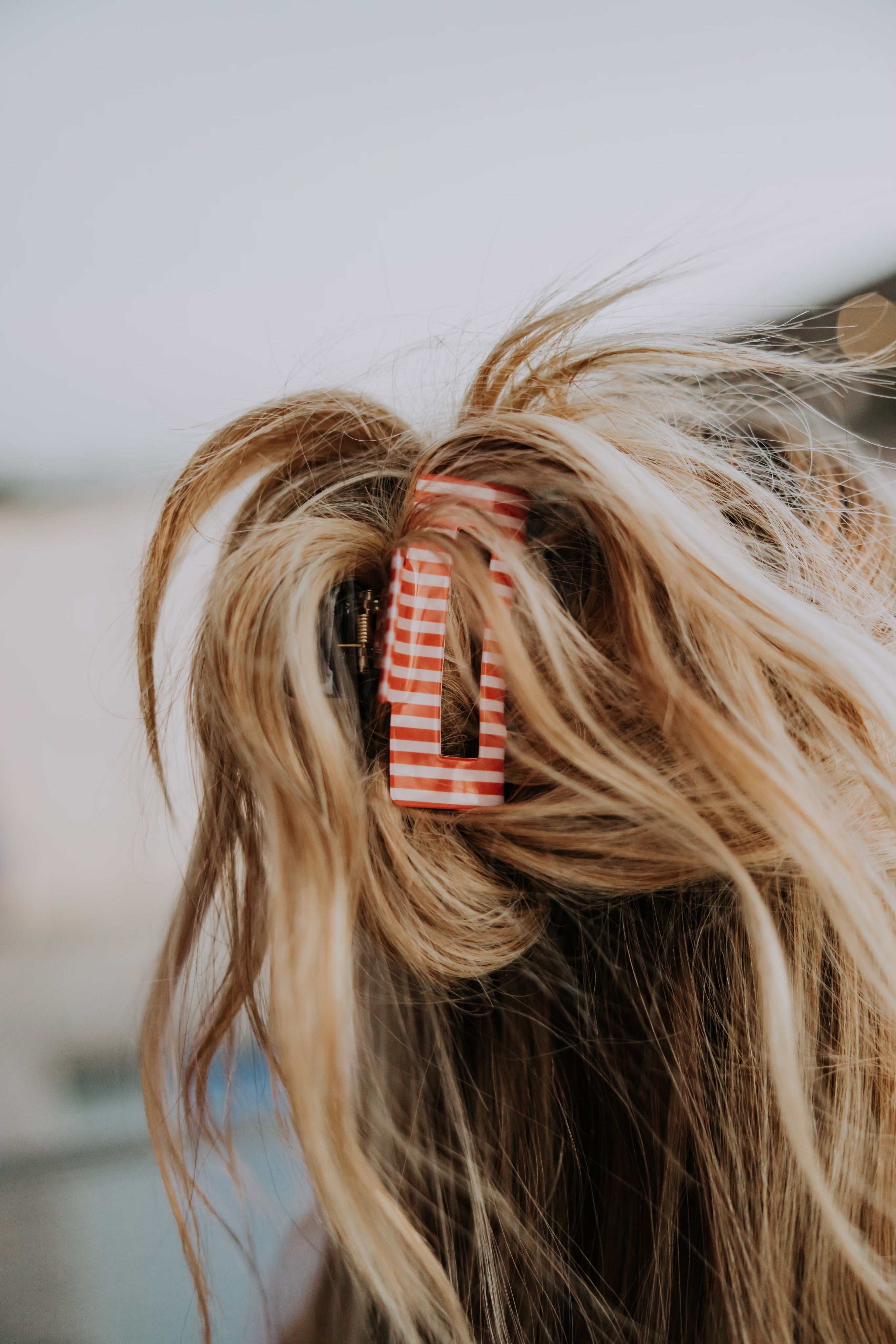 A close-up of messy blonde hair, secured with the Mama X™ Brand Red white and blue/Stripe Claw Clip, featuring red-and-white stripes. The clip is photographed from the back against a blurred background.