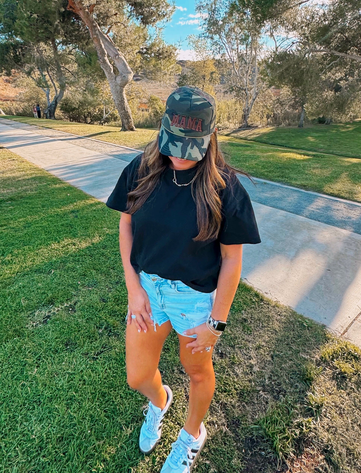 A woman wearing the Mama X™ Brand “Brown Camo Foam Trucker” hat, a black shirt, and denim shorts stands on grass near a sidewalk with one hand in her pocket, looking down. Trees and a clear sky are visible in the background.