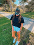 A woman wearing the Mama X™ Brand “Brown Camo Foam Trucker” hat, a black shirt, and denim shorts stands on grass near a sidewalk with one hand in her pocket, looking down. Trees and a clear sky are visible in the background.