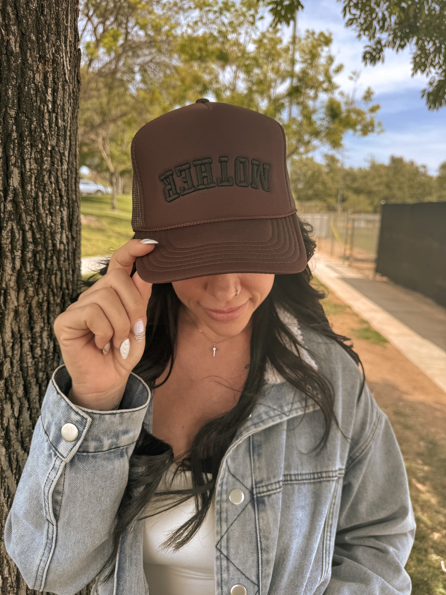 A woman outdoors tips a brown Mother Foam Trucker hat by Mama X™ Brand. With long dark hair, a light blue denim jacket, and white top, she stands beside a tree on a sunny day.