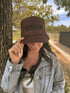 A woman outdoors tips a brown Mother Foam Trucker hat by Mama X™ Brand. With long dark hair, a light blue denim jacket, and white top, she stands beside a tree on a sunny day.
