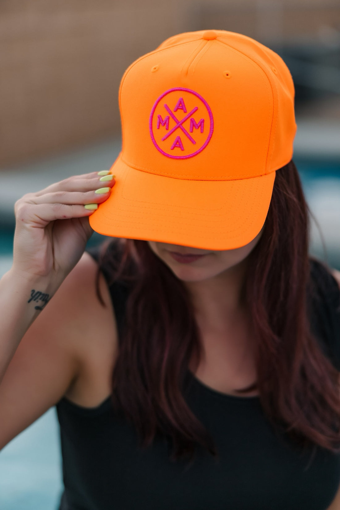A person with long brown hair tilts down a Neon Orange Mama X trucker hat by Mama X™ Brand, featuring a pink circular "MAMA" logo. They wear neon yellow-green nail polish and a black sleeveless top. The background is blurred.
