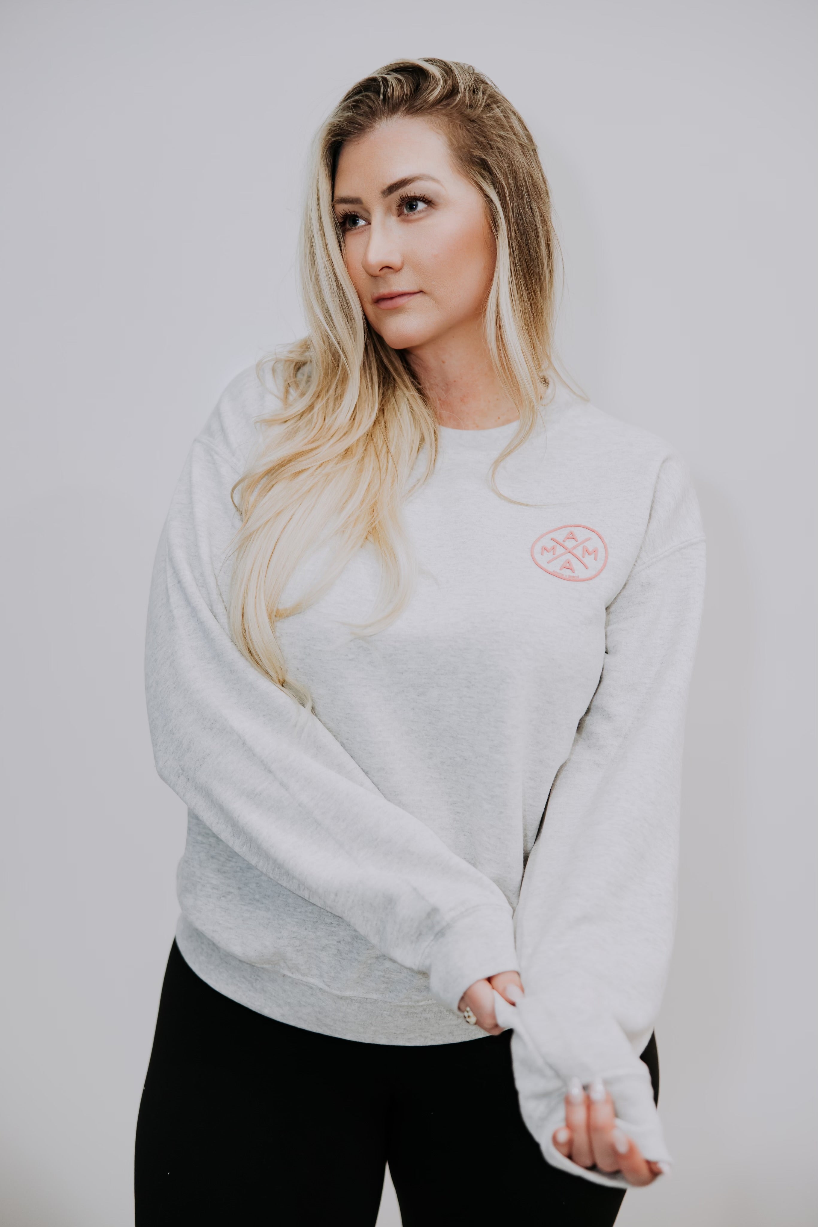 A woman with long blonde hair wears a Mama X™ Brand light gray crewneck and black pants, standing against a plain white background, looking to the side while holding the sleeves.