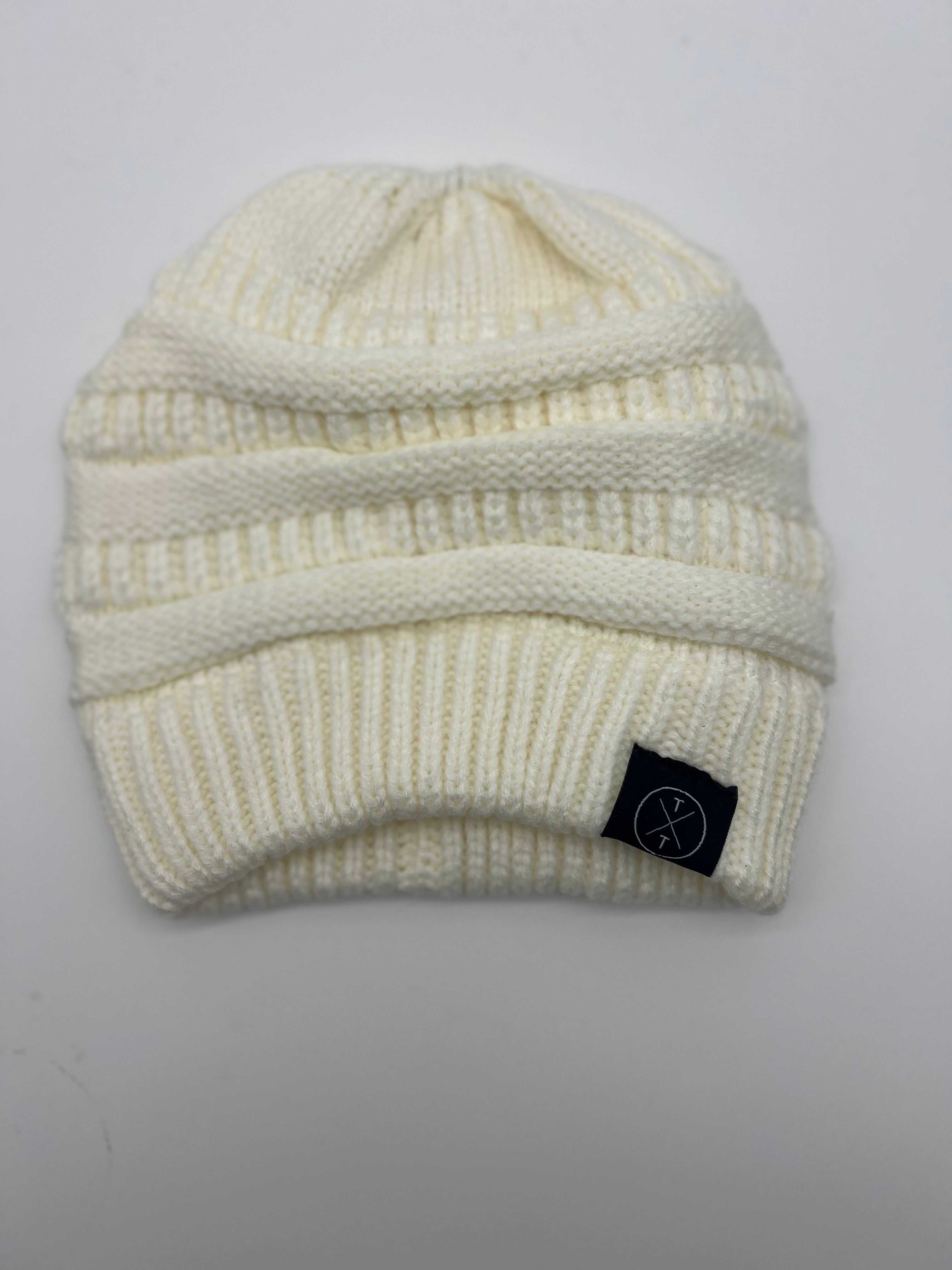 A white Knit Beanie for Adults with Ponytail Hole from Mama X™ Brand is displayed on a plain white background. The hat features a ribbed edge and folded cuff design. A small black tag with a minimalist logo is sewn on the left side of the cuff.