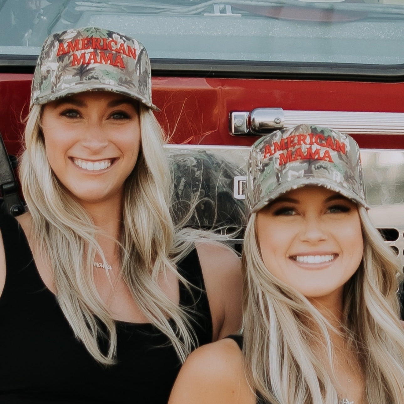 Two smiling women with long blonde hair wear Mama X™ Brand American Mama Hats, black tank tops, and denim shorts while sitting in front of a red truck. One holds a small American flag, showing their patriotic spirit.