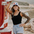 A smiling woman with tattoos, in a black tank, denim shorts, red cowboy boots, and the Red, White & Blue Mama Hat by Mama X™ Brand, poses outdoors near a red fire truck on a sunny day.