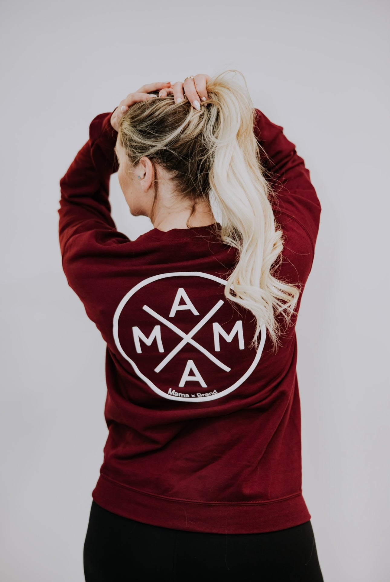 A person with long blonde hair, facing away, wears the Mama X™ Brand maroon crewneck. The design features "MAMA" in a circular textured puff print logo as they hold their hair in a ponytail.