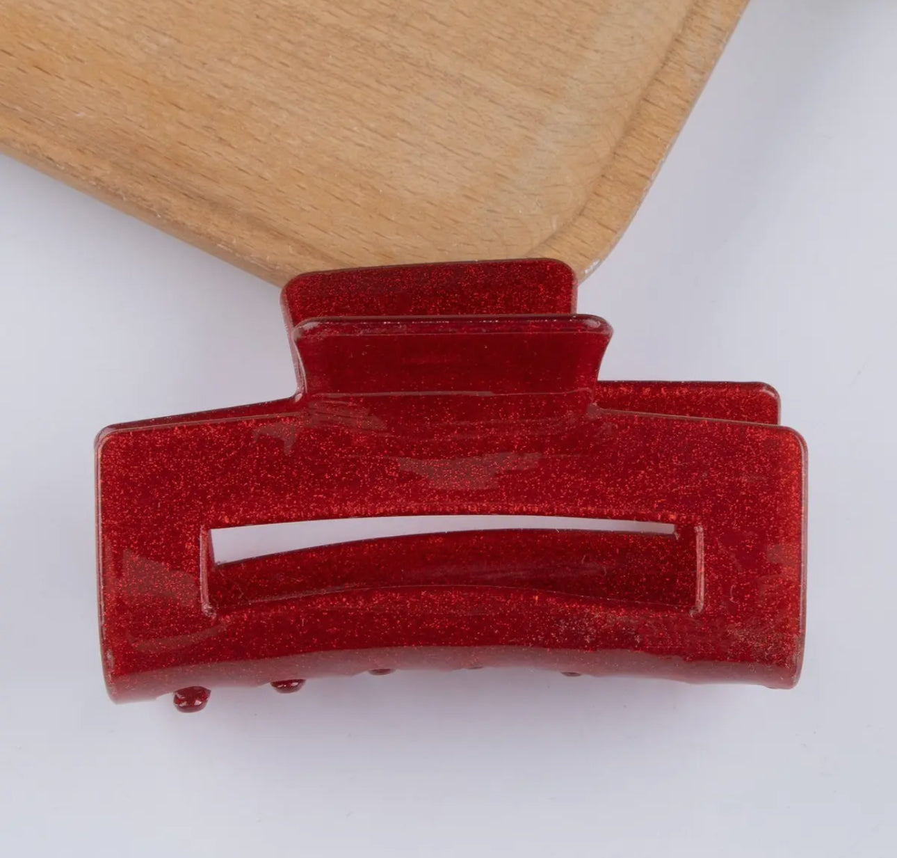 The Mama X™ Brand Red claw clip, crafted from durable acrylic with a glitter finish, sits on a light wooden surface against a white background, combining strong grip and stylish sparkle.