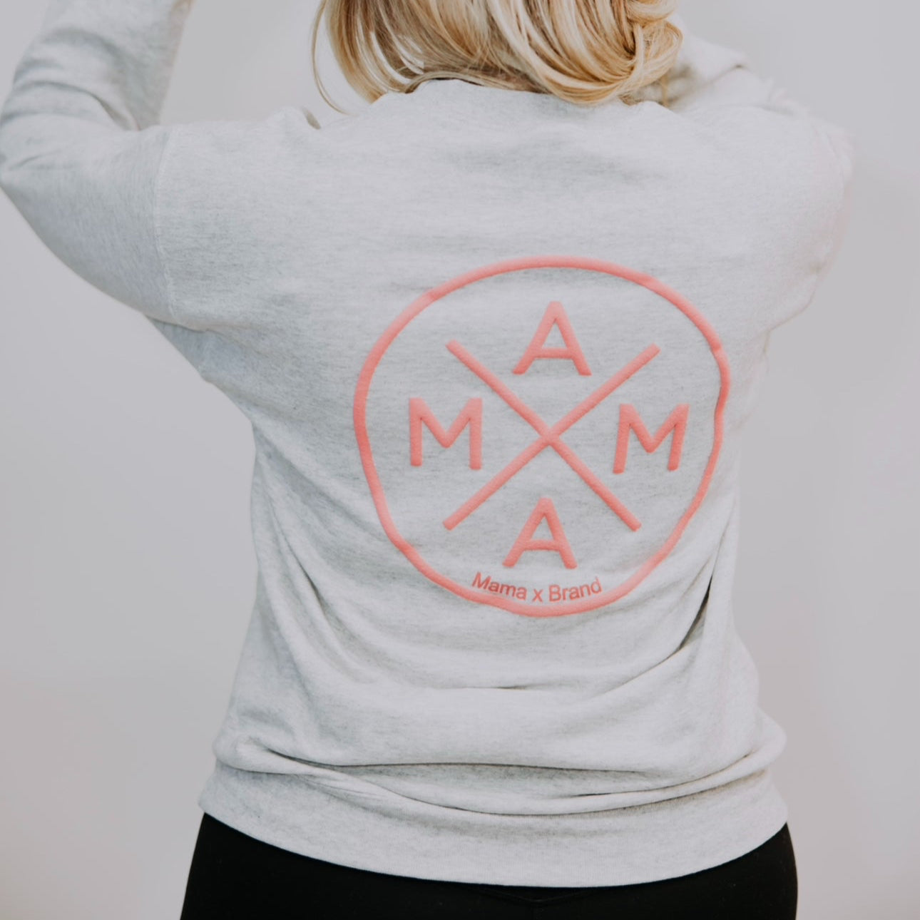 A person with blonde hair facing away wears the Mama X™ Brand Gray Crewneck, showcasing the relaxed fit "Mama X" logo in pink and white within a crossed circle design. They pair it with a matching baseball cap against a plain gray backdrop.