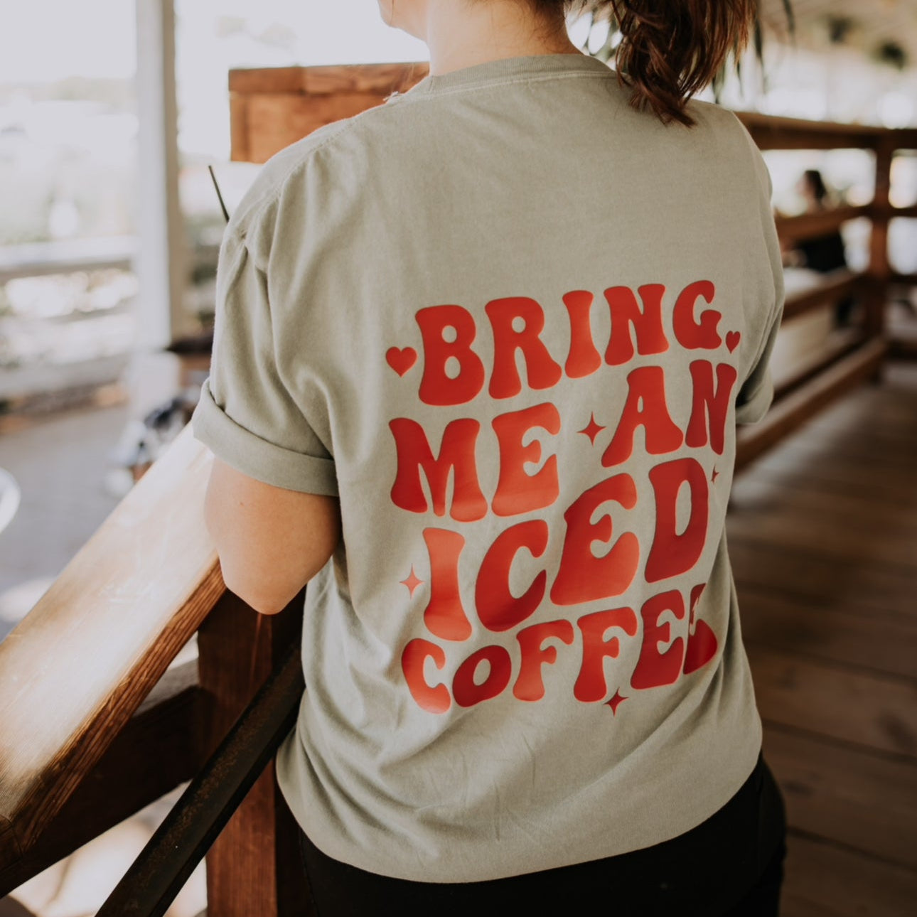 A woman with a ponytail faces away indoors, wearing the Bring Me An Iced Coffee Tshirt by Mama X™ Brand. The shirt features red "BRING ME AN ICED COFFEE" text and the Mama X™ logo, available now for preorder.