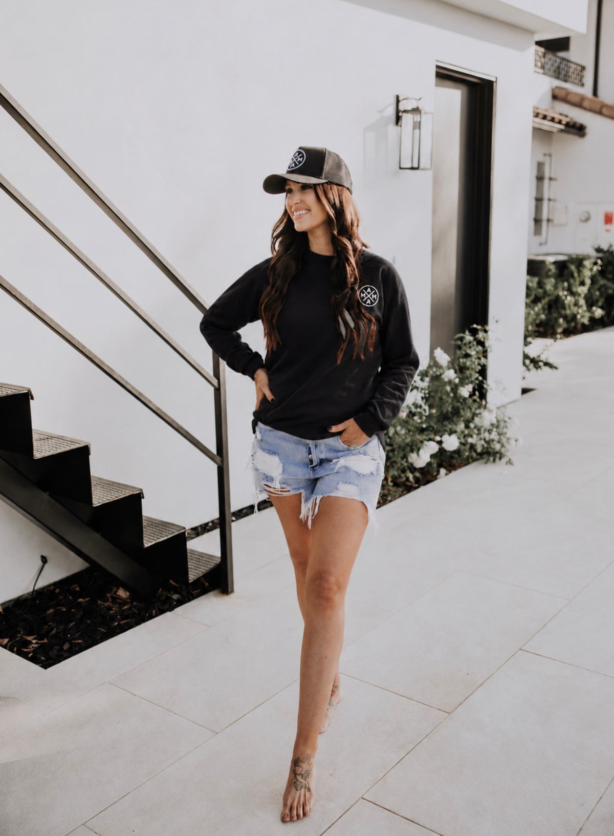A woman wearing a black Embroidered Mama X Unisex Sweatshirt with a small white circular logo on the chest, a black baseball cap, and distressed denim shorts.