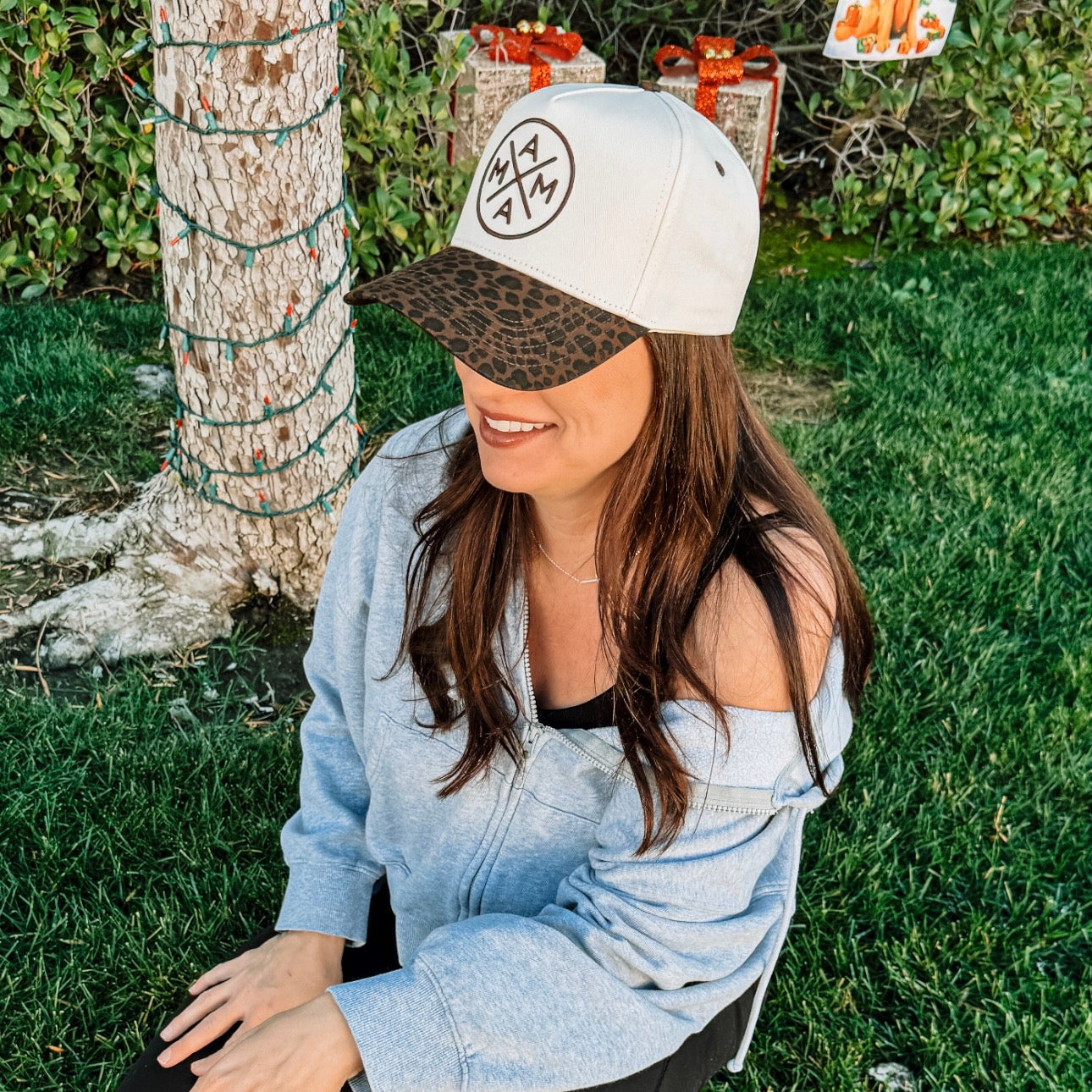 A woman wearing the Cheetah Mama X Trucker by Mama X™ Brand kneels on grass near a tree wrapped with lights, smiling to the side in a blue off-shoulder sweatshirt, black leggings, and slip-on shoes.
