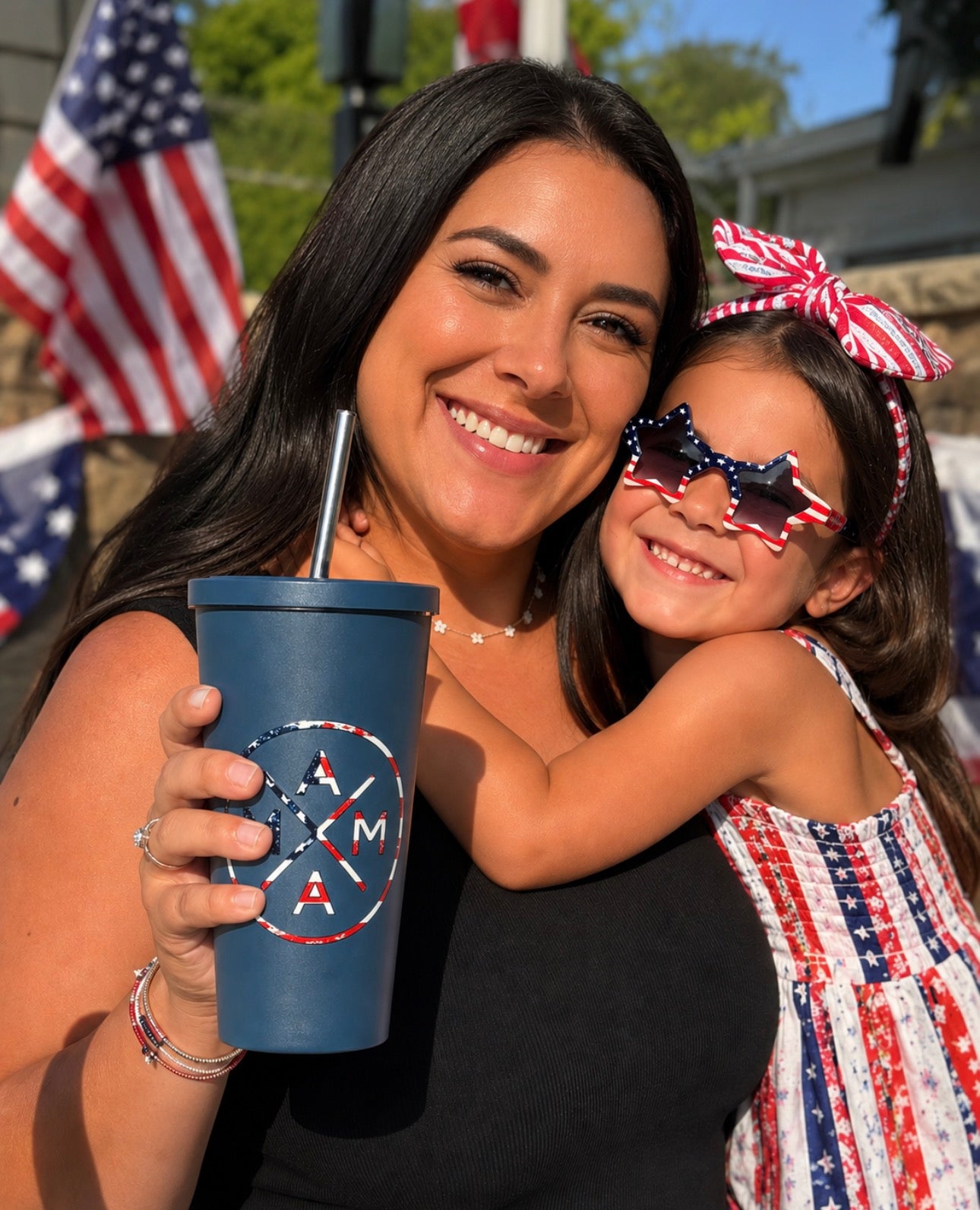 A smiling woman holds a Mama X™ Brand Mama X American Flag Tumbler and hugs a young girl in star-shaped American flag sunglasses and a patriotic dress, with U.S. flags and festive decorations in the background.
