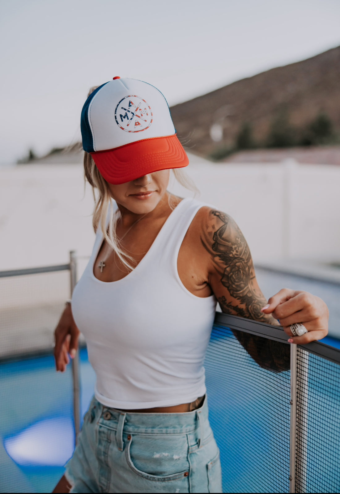 A woman in a white tank, denim shorts, and the American Flag Mama X™ Foam Trucker Hat by Mama X™ Brand leans on a metal fence by a pool. She has a tattoo sleeve, necklace, and rings. Mountains are visible in the blurred background.