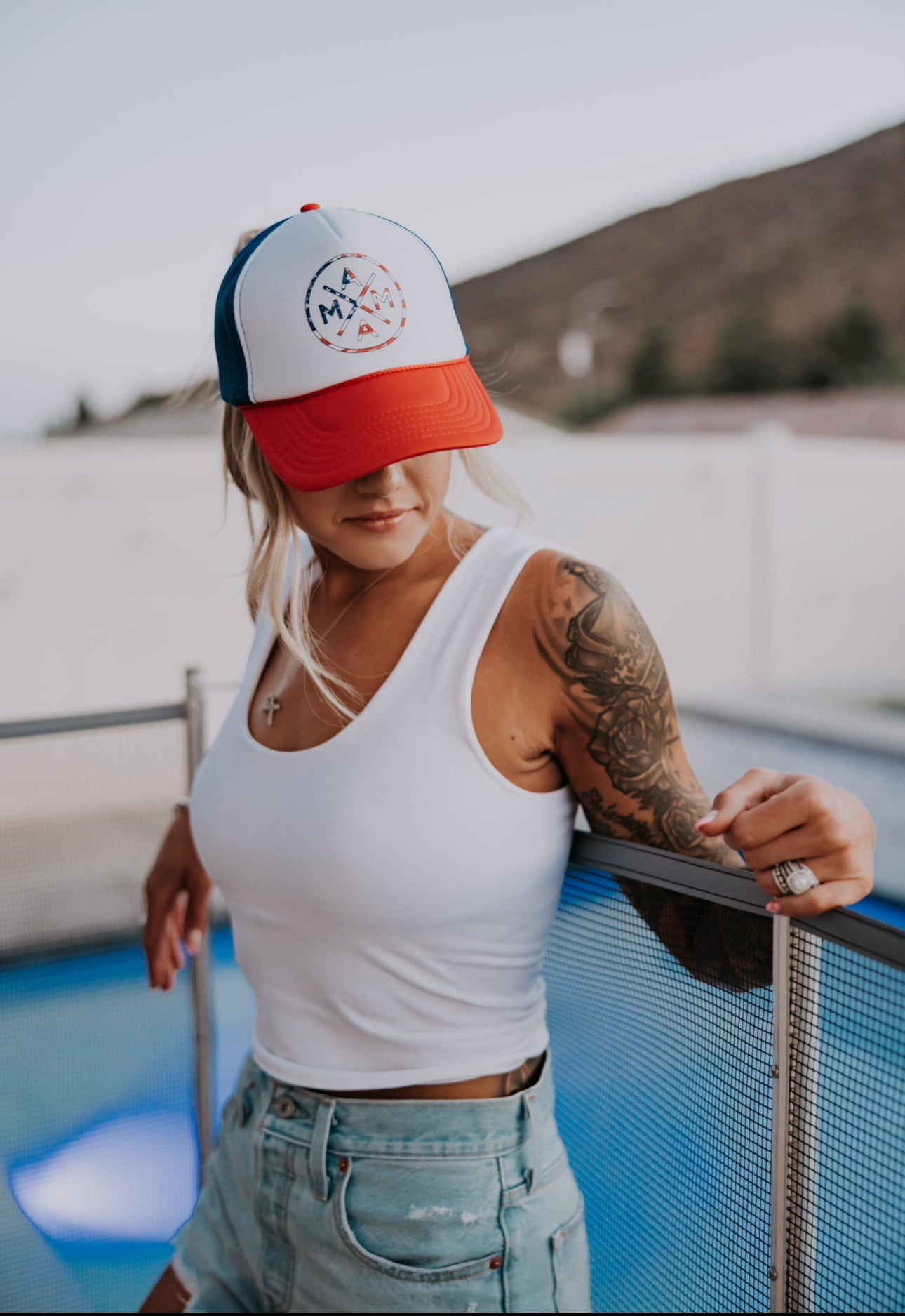 A woman in a white tank, denim shorts, and the American Flag Mama X™ Foam Trucker Hat by Mama X™ Brand leans on a metal fence by a pool. She has a tattoo sleeve, necklace, and rings. Mountains are visible in the blurred background.