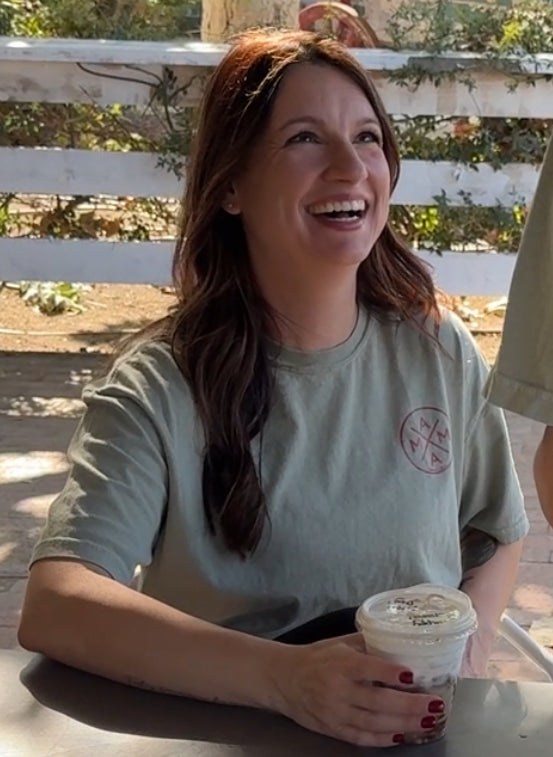 A woman with long brown hair smiles outdoors, holding an iced coffee and wearing the "Bring me an iced coffee" T-shirt by Mama X™ Brand as sunlight shines behind her.