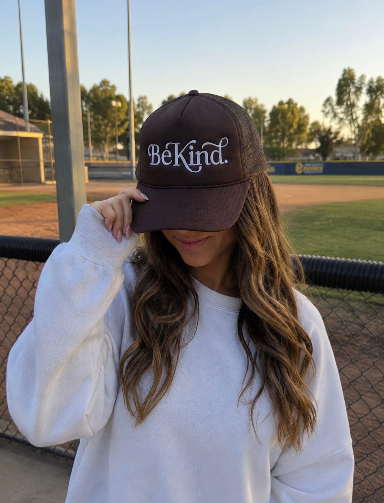 Wearing the Mama X™ Brand “Be Kind Trucker” cap and a white sweatshirt, a person with long wavy hair stands by a baseball field, partially covering their face with the hat.