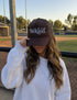 Wearing the Mama X™ Brand “Be Kind Trucker” cap and a white sweatshirt, a person with long wavy hair stands by a baseball field, partially covering their face with the hat.