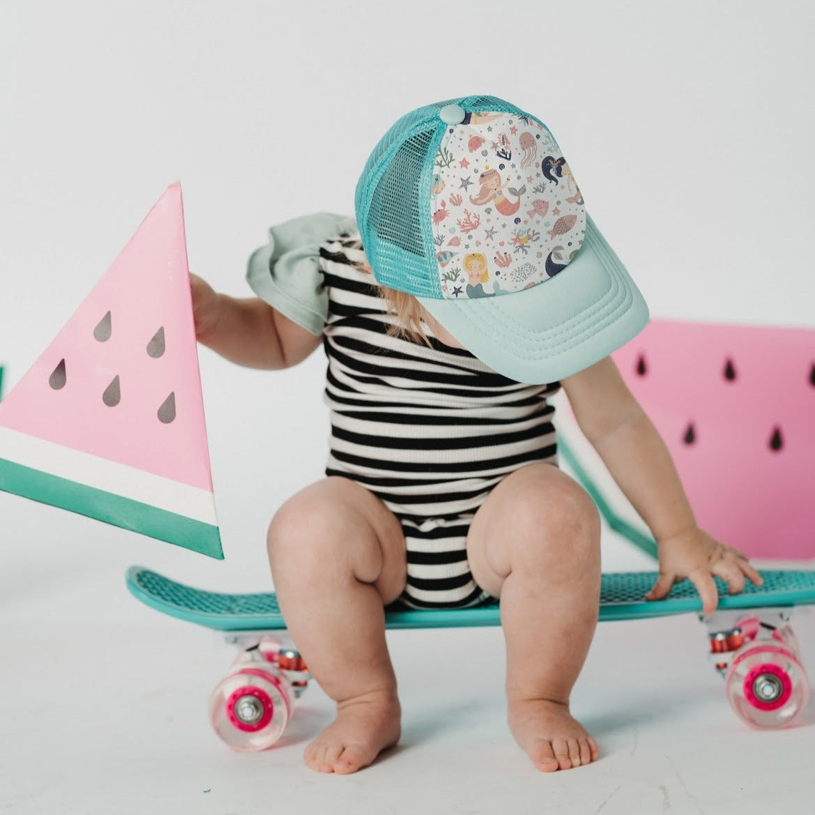 A baby wearing a turquoise Mermaid Trucker Hat from Mama X™ Brand adorned with playful sea creatures and a black-and-white striped onesie sits on a turquoise skateboard. The baby holds a pink toy watermelon slice with a green rind. The background features a white wall with pink accents, bringing an enchanting touch of unicorns to the scene.