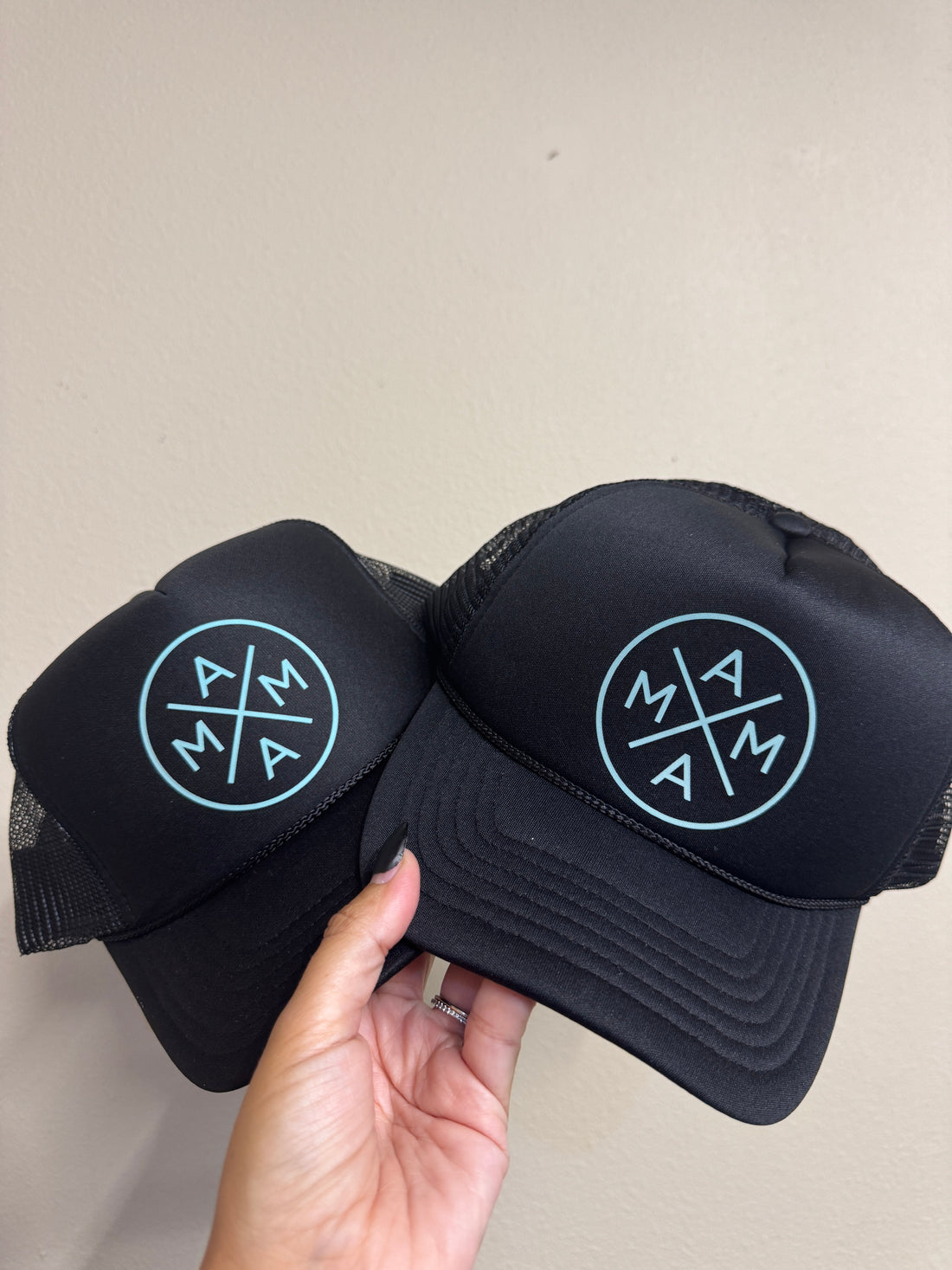 A hand holds two Black foam truckers with teal logos from Mama X™ Brand, featuring “MAMA” letters arranged around an "X" on the front. The background is a plain beige wall.