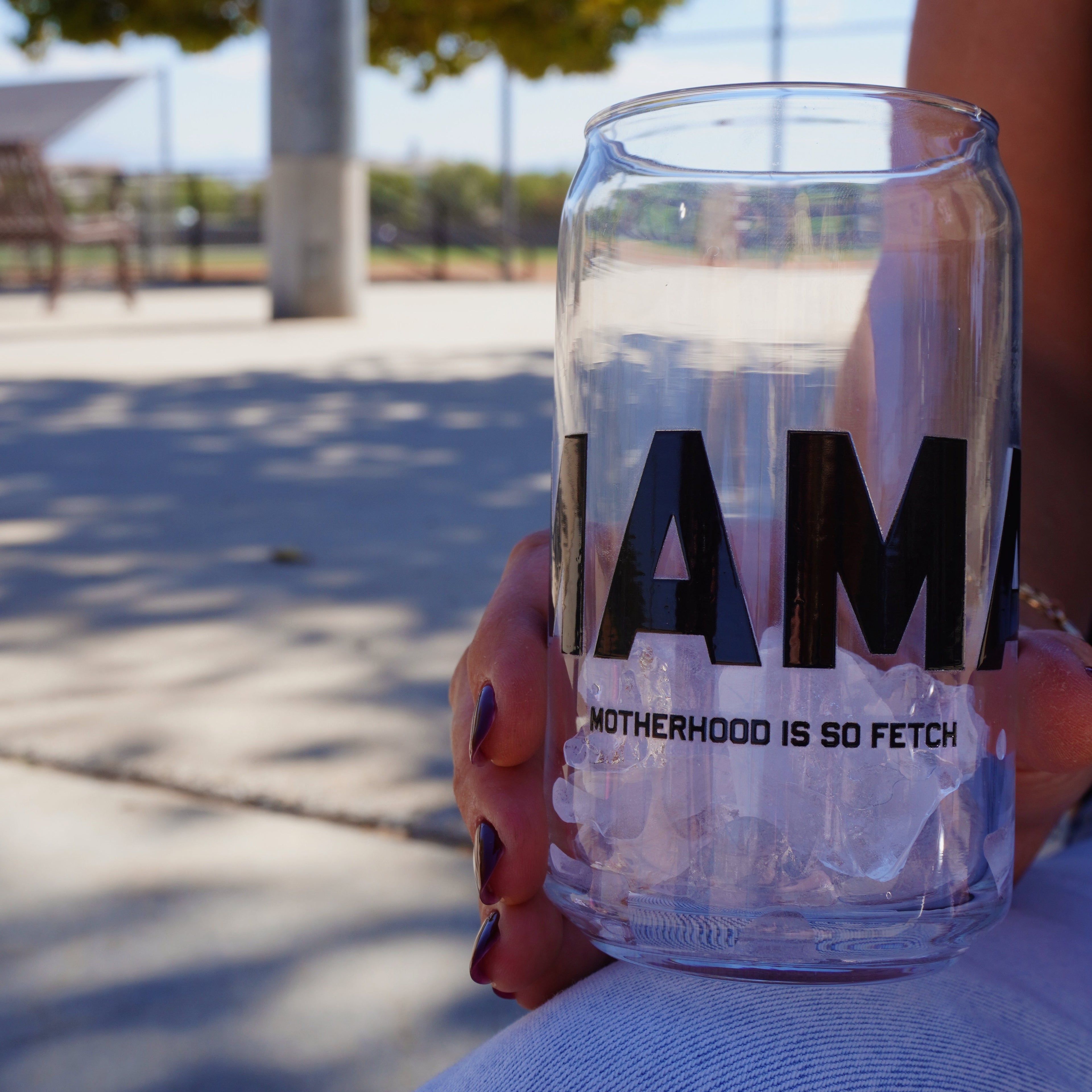 A person with painted nails holds a 16 oz clear Motherhood Is So Fetch Cup by Mama X™ Brand—printed with "MAMA" and "MOTHERHOOD IS SO FETCH"—filled with ice, while relaxing outdoors on a sunny day.