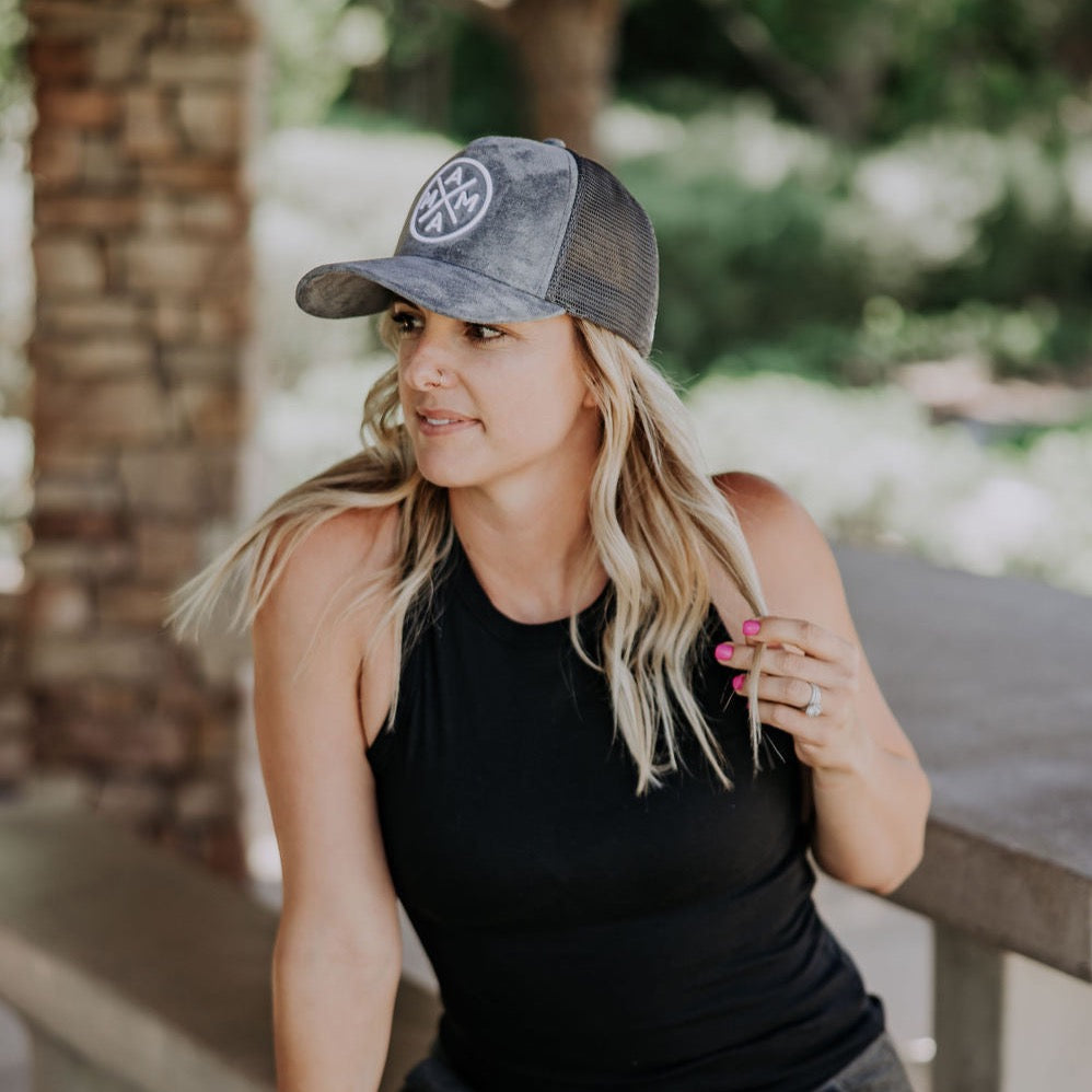 A woman with long blonde hair wearing a black sleeveless top and a Mama X™ Trucker Hat in gray suede is standing outside. She is holding a strand of her hair with one hand and has a slight smile on her face. The background is blurred, showing greenery and a stone pillar.