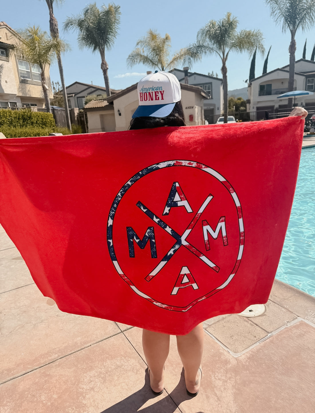 By the pool, someone shows off the Mama x American Flag Towel by Mama X™ Brand, pairing it with an "American Honey" cap. Palm trees and houses frame a clear sky—an ideal setting to celebrate with this 4th of July towel.