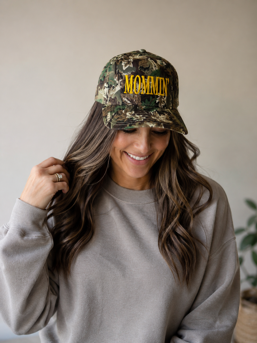 A woman with long wavy hair smiles while wearing the Mama X™ Brand "Mommin camo in yellow" hat and a beige sweatshirt, looking down as she touches her hair.