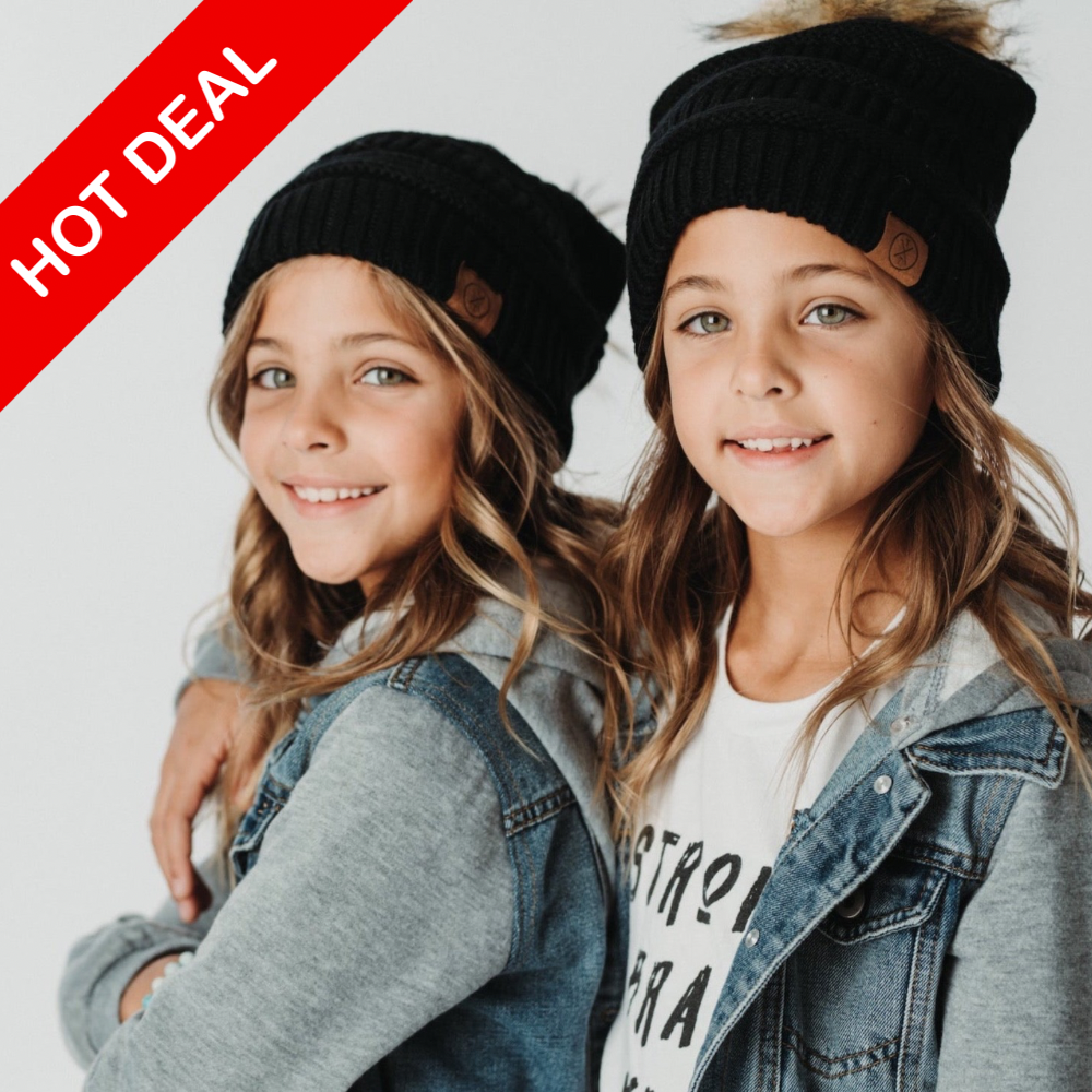 Two cheerful children wearing Child Pom Beanies in black from Mama X Brand and denim jackets stand together. A red label with white text reads 