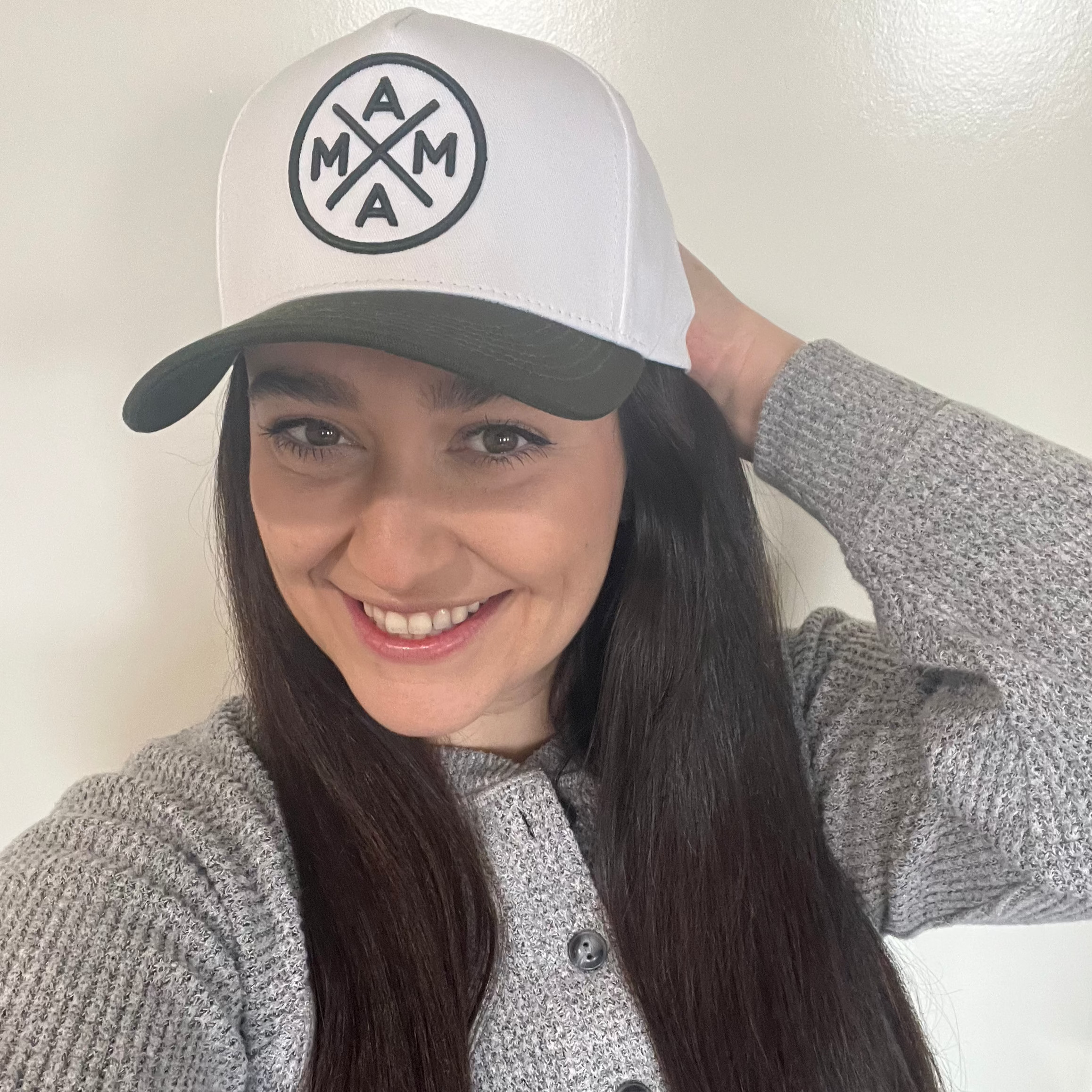 A person with long dark hair smiles while wearing a light gray sweater and the Dark Green Mama X™ Baseball Cap, emblazoned with the iconic circular logo from Mama X™ Brand, perfect for the holidays. They are posing indoors against a light-colored wall.