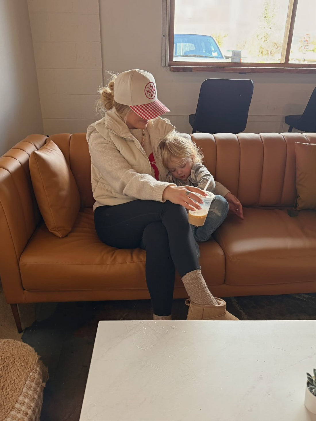 A woman wearing the Mama X™ Brand Red Gingham Mama X Trucker hat sits on a tan sofa with a child, enjoying a drink together. They look cozy and relaxed in a bright room with chairs and a window in the background.