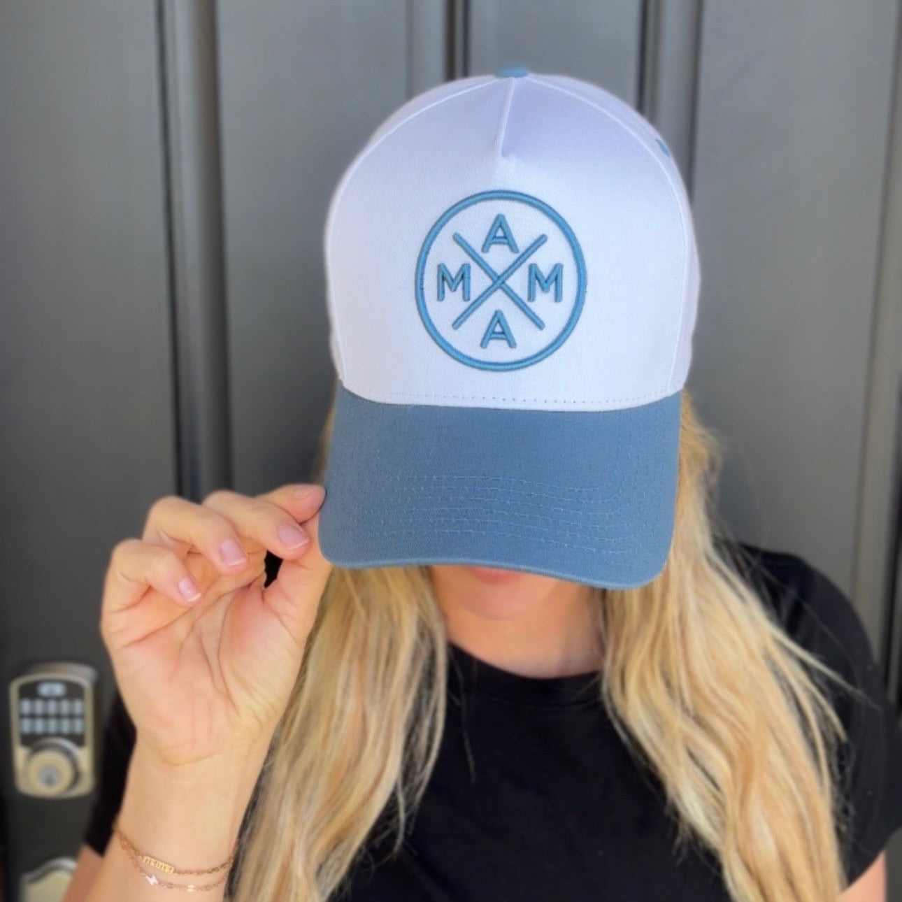 A long-haired person with blonde hair, wearing a black shirt and the Blue Mama X™ Baseball Cap from Mama X™ Brand, is standing in front of a gray door, slightly tipping their cap.