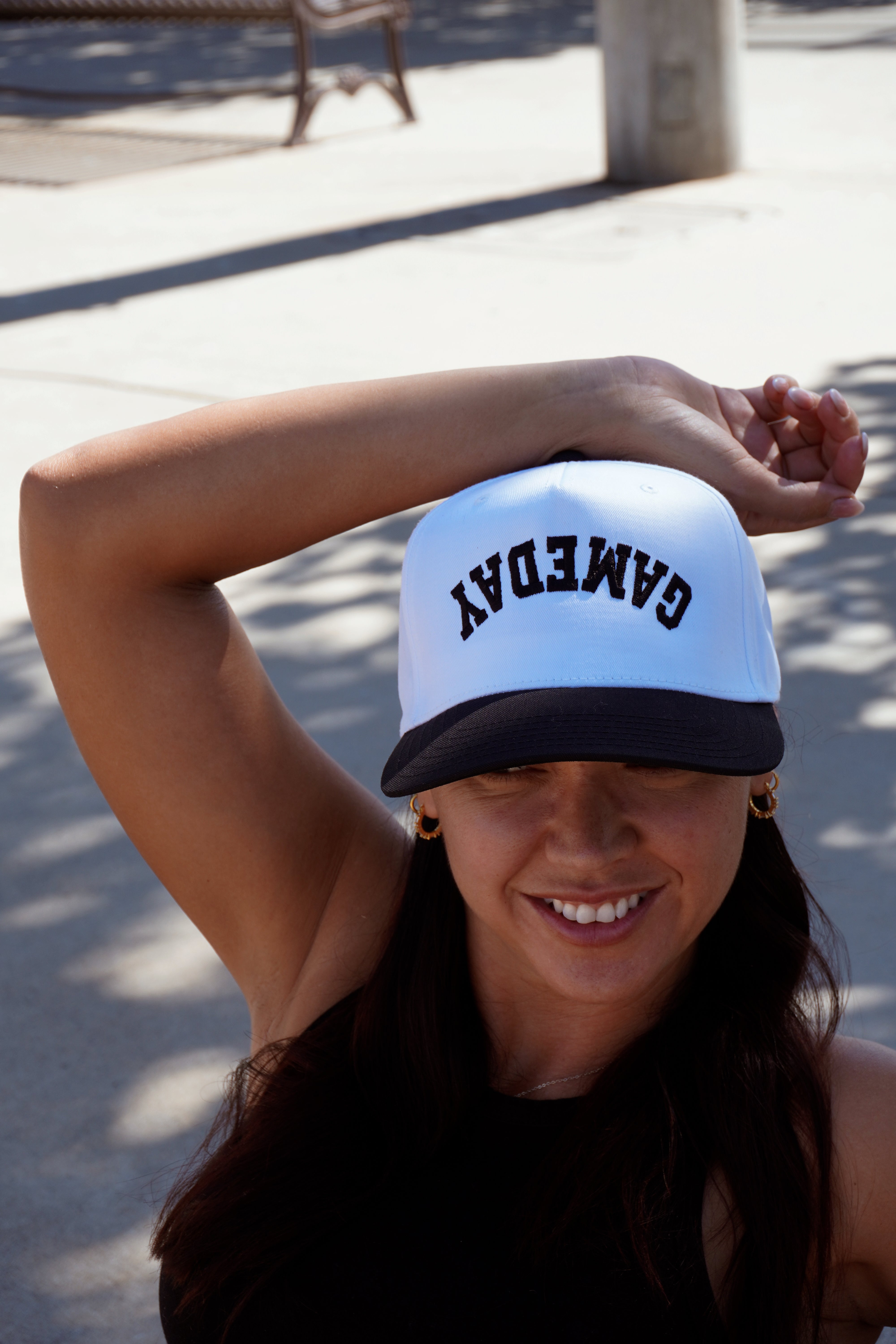 A smiling woman outdoors on a sunny day poses with one arm raised, wearing the Mama X™ Brand “Game Day Trucker” cap with “GAME DAY” written upside down in black and white.