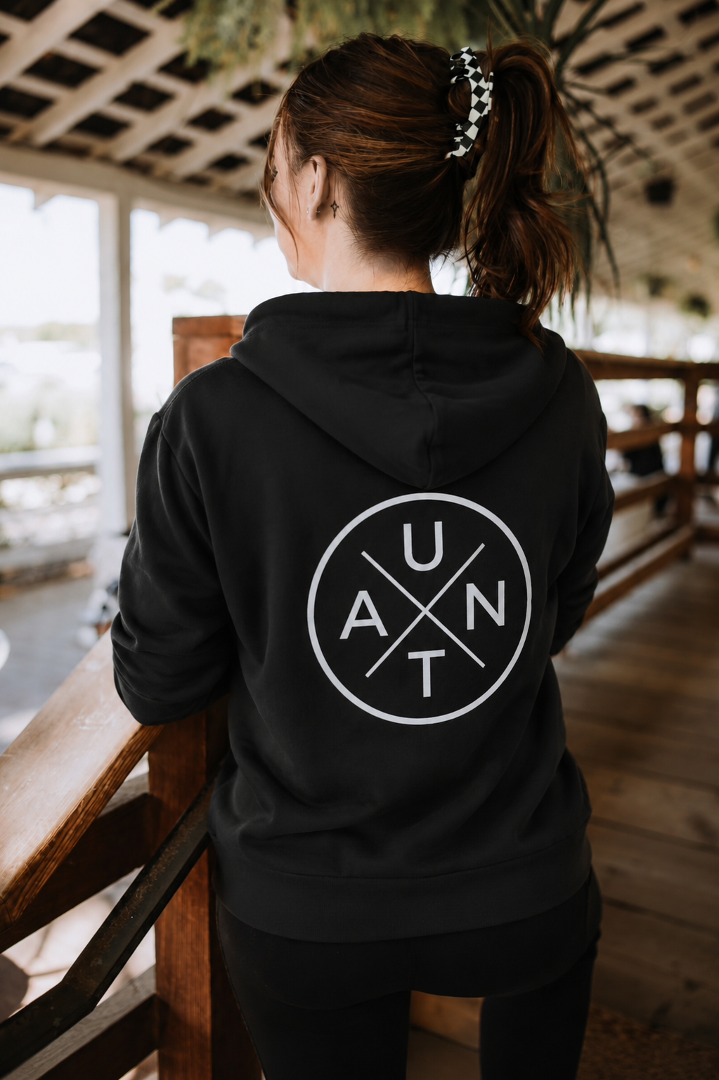 A woman with a ponytail wears a black Mama X™ Brand Aunt fleece zip up hoodie, perfect for cozy comfort and everyday wear, standing indoors by a wooden railing—an ideal aunt gift.