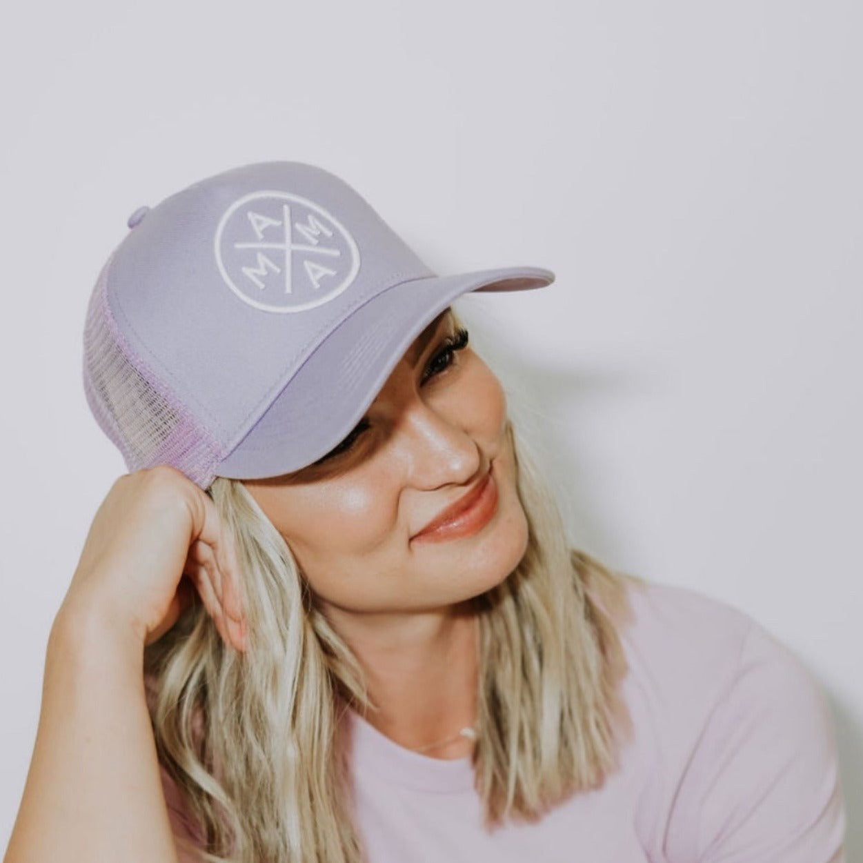 A person with blond hair, wearing a Mama X™ Premium Canvas Trucker Hat in Lavender from the Mama X™ Brand, is smiling and looking to the side. The person is also wearing a light pink shirt and has their head tilted slightly, with one hand resting on the brim of the cap.