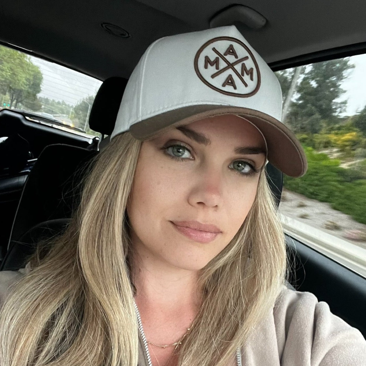 A person with long blonde hair is sitting in a car wearing a Brown Mama X™ Baseball Cap by Mama X™ Brand, featuring a circular logo. They are looking at the camera with a neutral expression. Trees, greenery, and a road can be seen through the car's windows in the background.