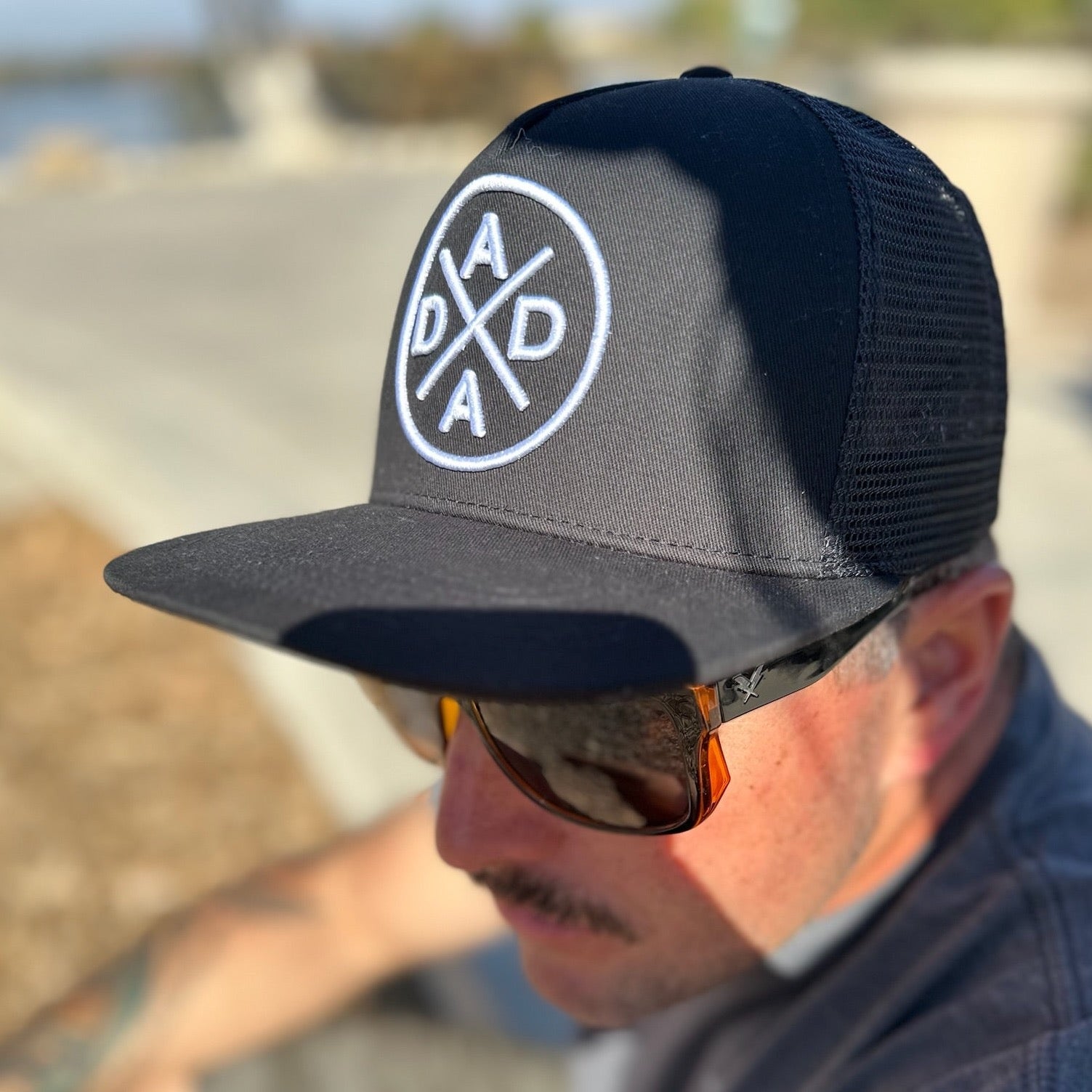 A person wearing a Dada X™ Premium Trucker Hat-Stitched 3D Logo from Mama X™ Brand with the logo embroidered on the front in a circular design sits outdoors. They have a mustache and are wearing brown sunglasses, with a blurred outdoor background visible behind them.