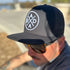 A person wearing a Dada X™ Premium Trucker Hat-Stitched 3D Logo from Mama X™ Brand with the logo embroidered on the front in a circular design sits outdoors. They have a mustache and are wearing brown sunglasses, with a blurred outdoor background visible behind them.