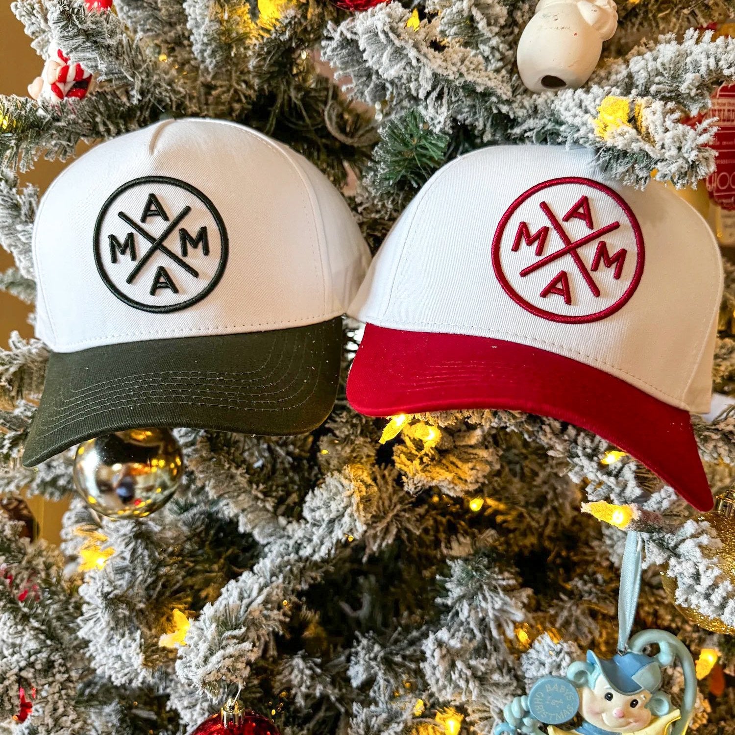 Two baseball caps from the Holiday Baseball Cap Bundle by Mama X™ Brand hang on a snow-covered Christmas tree. The cap on the left features a white front with a green brim and a black logo, while the cap on the right sports a white front with a red brim and red logo. The tree is beautifully adorned with lights and ornaments.