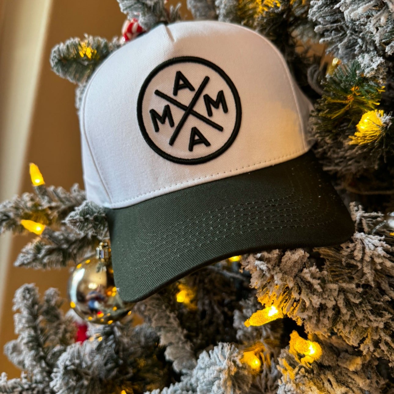 A Dark Green Mama X™ Baseball Cap hangs on a Christmas tree, adorned with warm yellow lights and frosted branches. Red and silver ornaments are visible in the background.