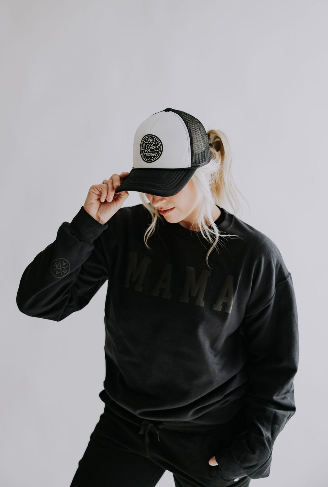 A person wearing a black "MAMA" sweatshirt is looking down and holding the brim of their Be A Nice Human Trucker Hat by Mama X™ Brand, against a light gray background.