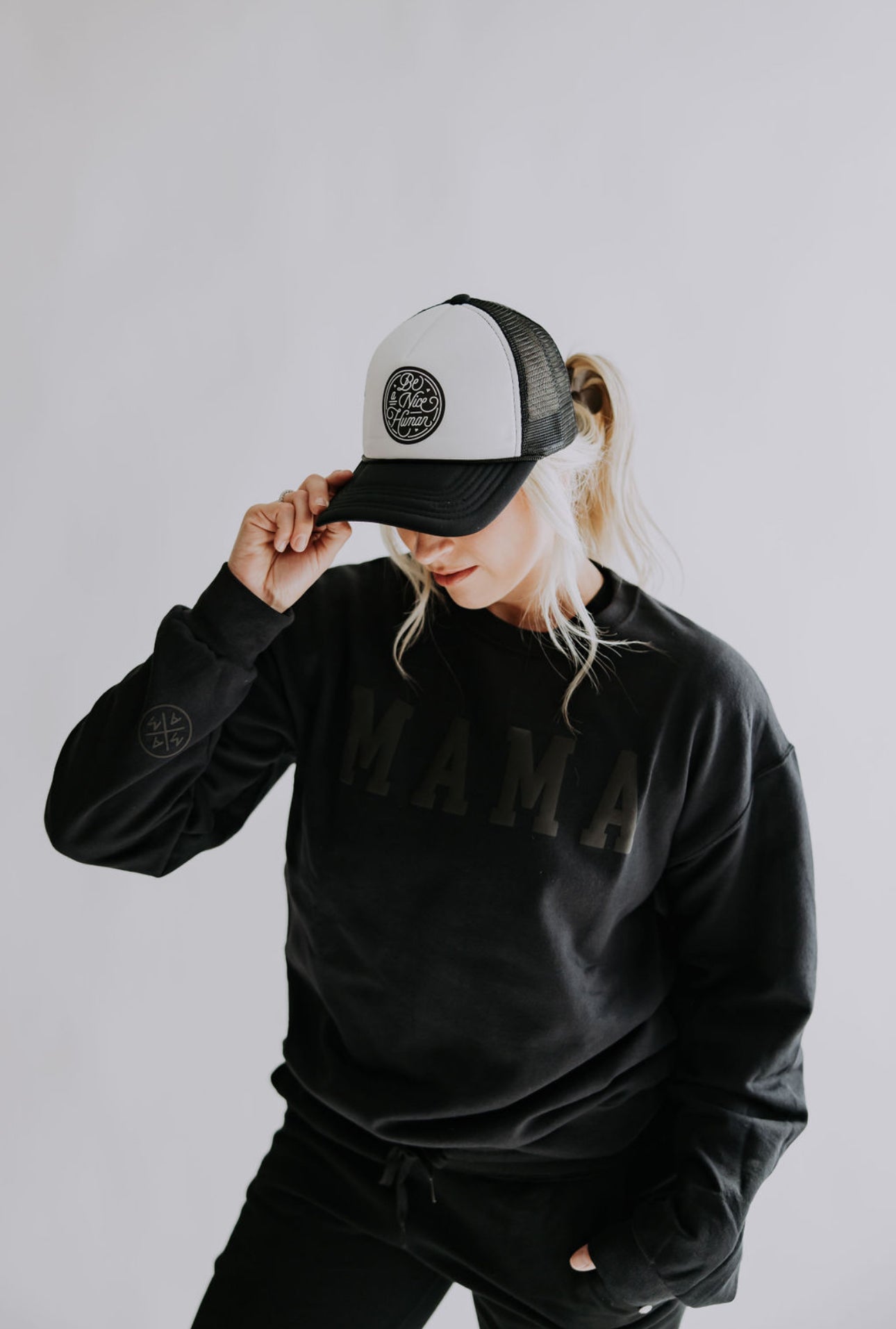 A person wearing a black "MAMA" sweatshirt is looking down and holding the brim of their Be A Nice Human Trucker Hat by Mama X™ Brand, against a light gray background.