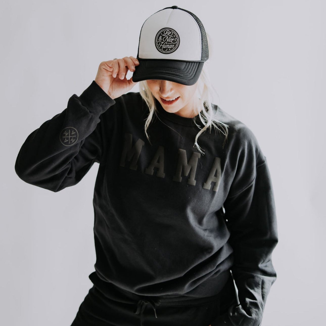 A woman wearing a black sweatshirt with "MAMA" printed on it, paired with the Be A Nice Human Trucker Hat from Mama X™ Brand, tilting her head slightly downward against a plain, light-colored background.