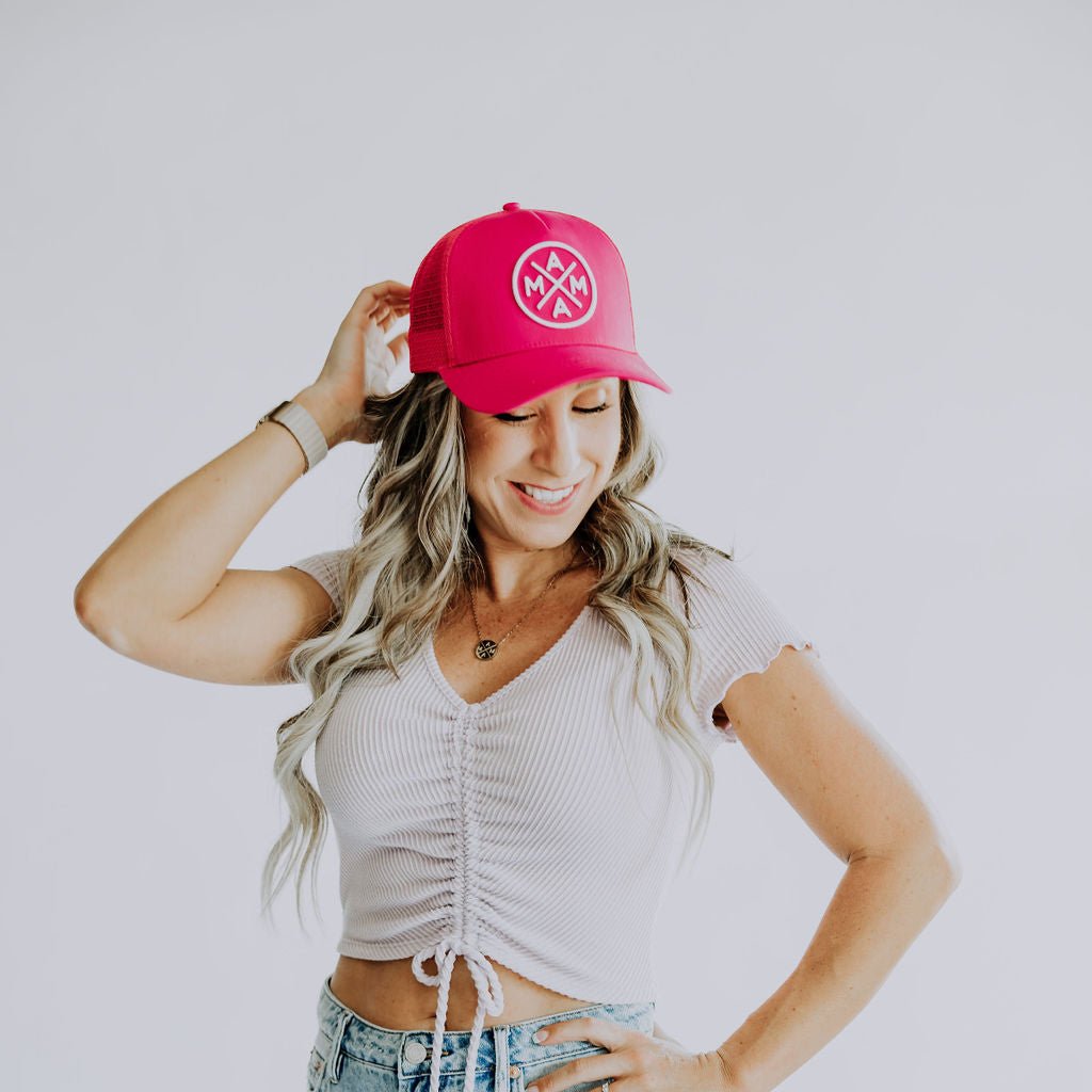 A woman with long wavy blonde hair wearing a Mama X™ Premium Canvas Trucker Hat in hot pink, a ruched white cropped top, and blue jeans is posing against a plain white background. She is smiling and holding the brim of her hat with one hand.